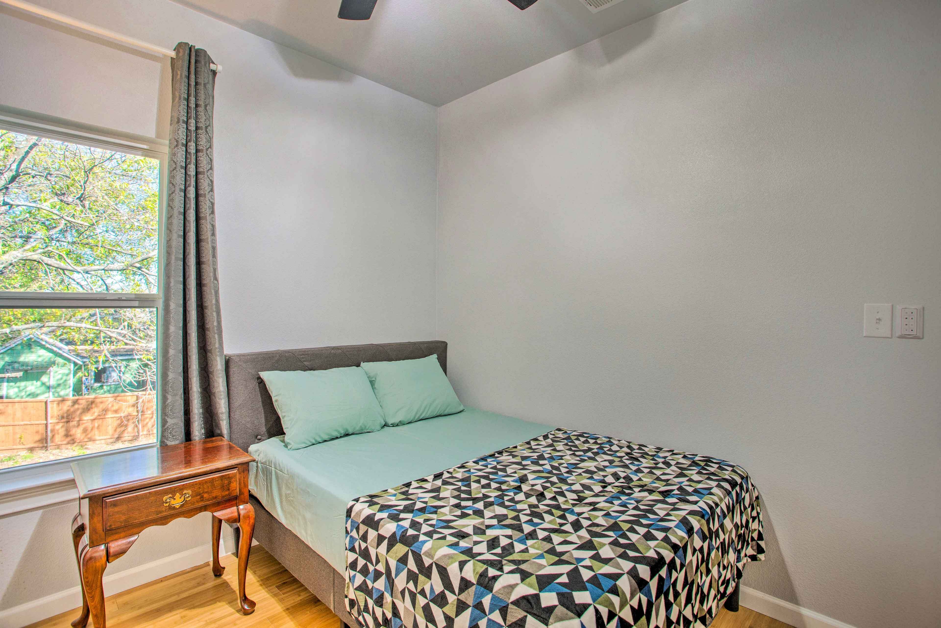 A bedroom with a bed, a nightstand, a window with curtains, and a view of greenery outside.