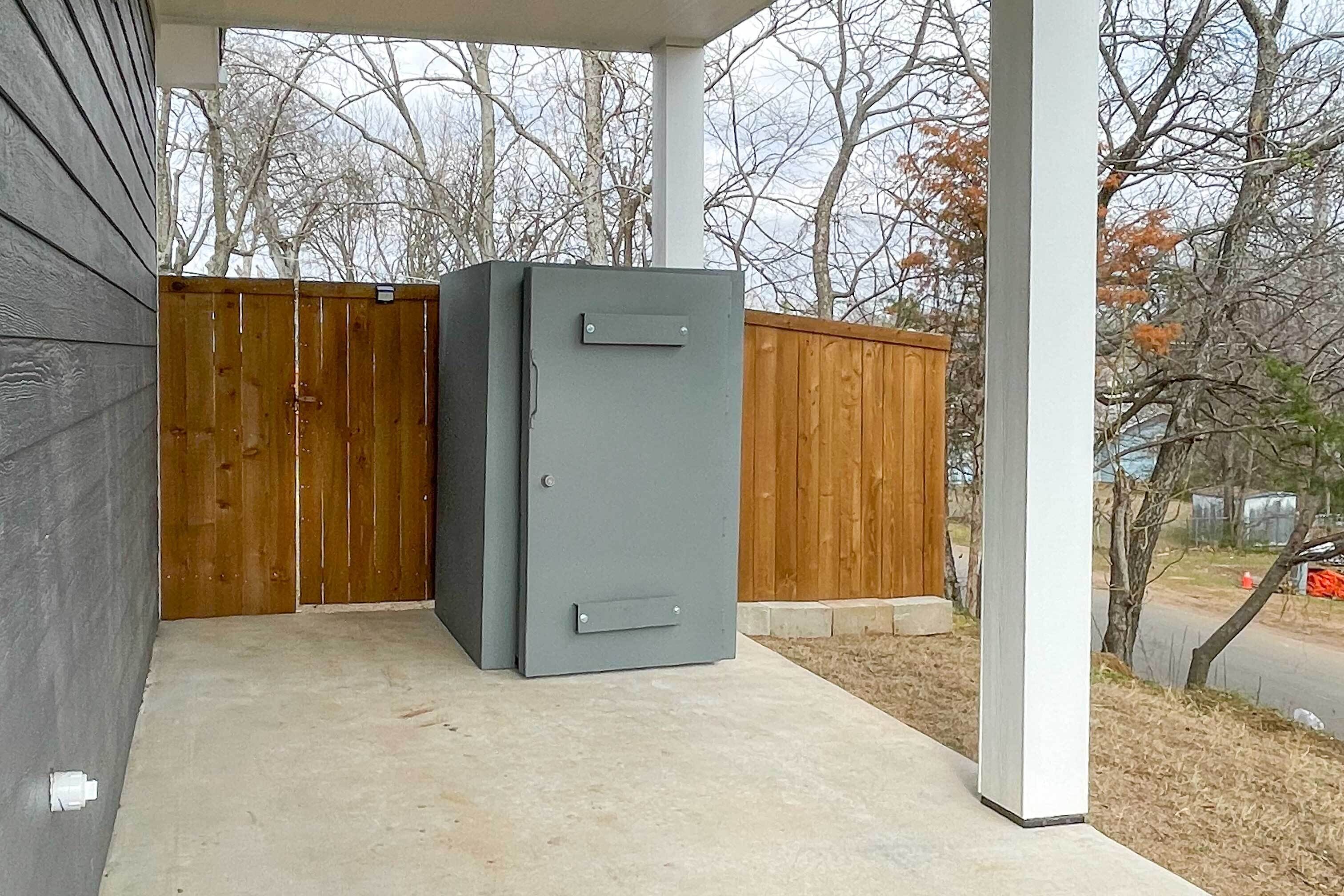 Una caja fuerte metálica gris en un espacio exterior cubierto, con un cerco de madera y árboles al fondo.