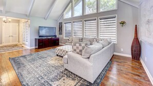 Ein geräumiges Wohnzimmer mit hoher Decke, einem großen Fenster mit Jalousien, einem grauen Sofa, einem Fernseher und einer dekorativen Vase.