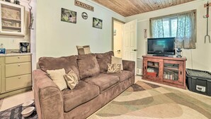 A cozy living room with a brown sofa, a television on a wooden cabinet, and a kitchen area with a fridge and a window with curtains. - Private Murray Abode w/ Fire Pit: Hunt & Fish (Murray)