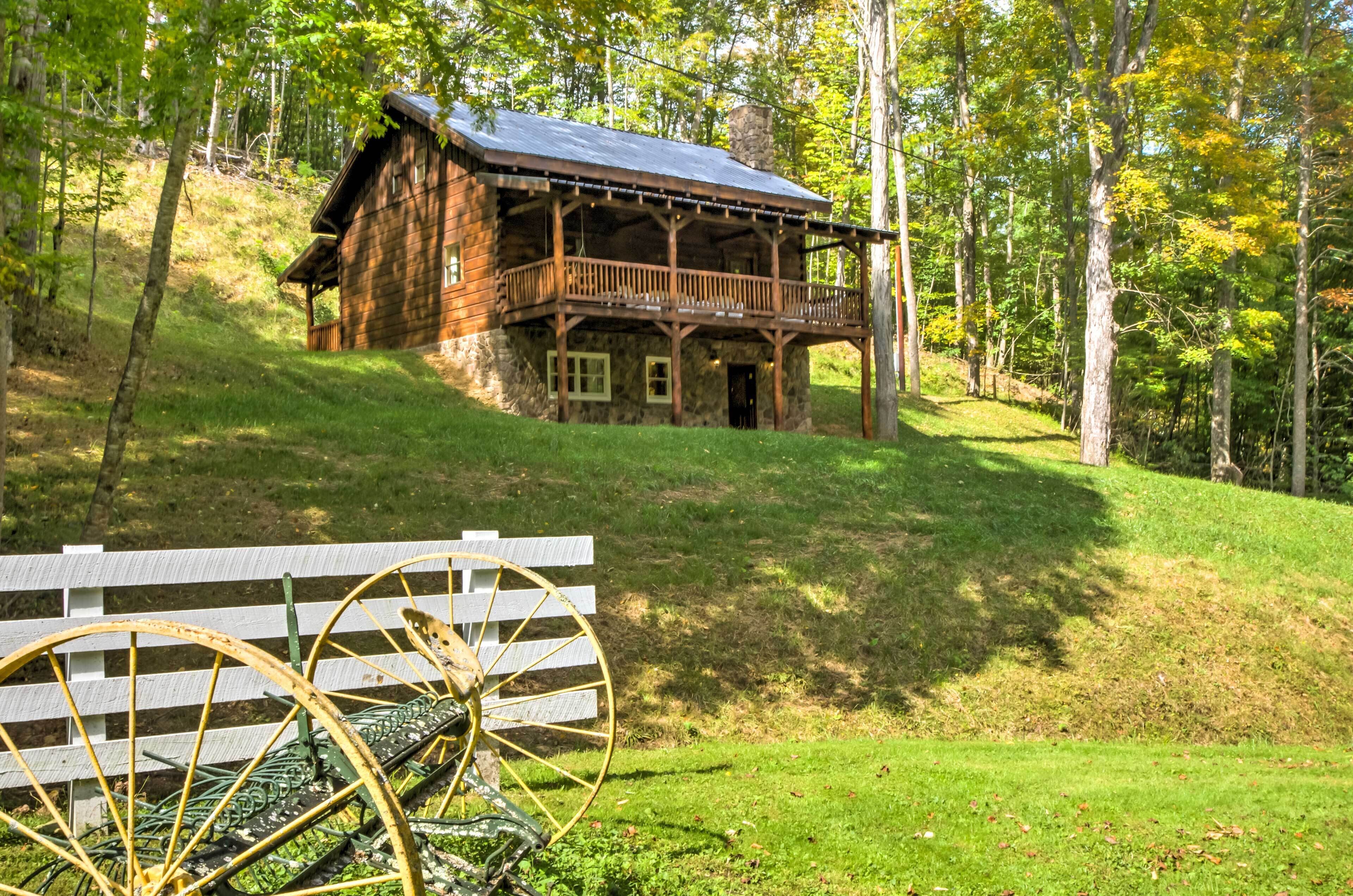 A rustic log cabin with a balcony, surrounded by a grassy hillside and trees.