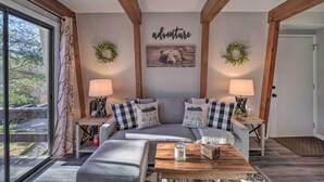 Un salon chaleureux avec un canapé gris, deux tables d’appoint avec des lampes, une table basse avec une bougie, et un mur orné d’une image d’ours et du mot « aventure ».