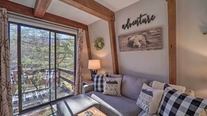 Un salon chaleureux avec une grande fenêtre, un plafond en bois, un canapé agrémenté de coussins à motifs et un mur orné d’une image d’ours et du mot « aventure ».