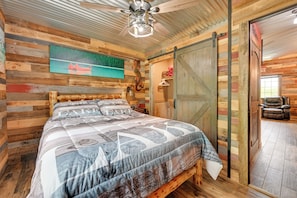 Une chambre rustique en bois, avec un lit, un ventilateur de plafond, une porte coulissante de style grange et une vue sur une chaise dans la pièce adjacente.