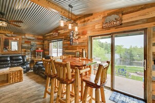 A rustic cabin interior with a wooden dining table, chairs, a black leather couch, and a large sliding glass door leading to a view of a lake.