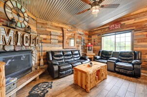 A rustic cabin interior with a fireplace, leather sofas, a wooden coffee table, and a large clock on the wall.