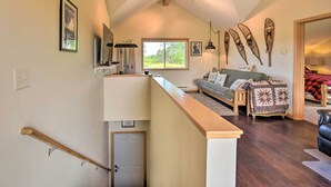 A spacious living area with a high ceiling, a wooden staircase, and a large window offering a view of greenery.