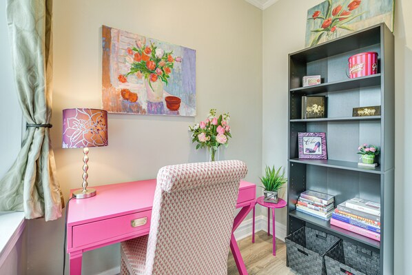 Un escritorio rosa con una lámpara, una estantería con libros y objetos decorativos, un cuadro de flores y una pequeña planta sobre una mesita.