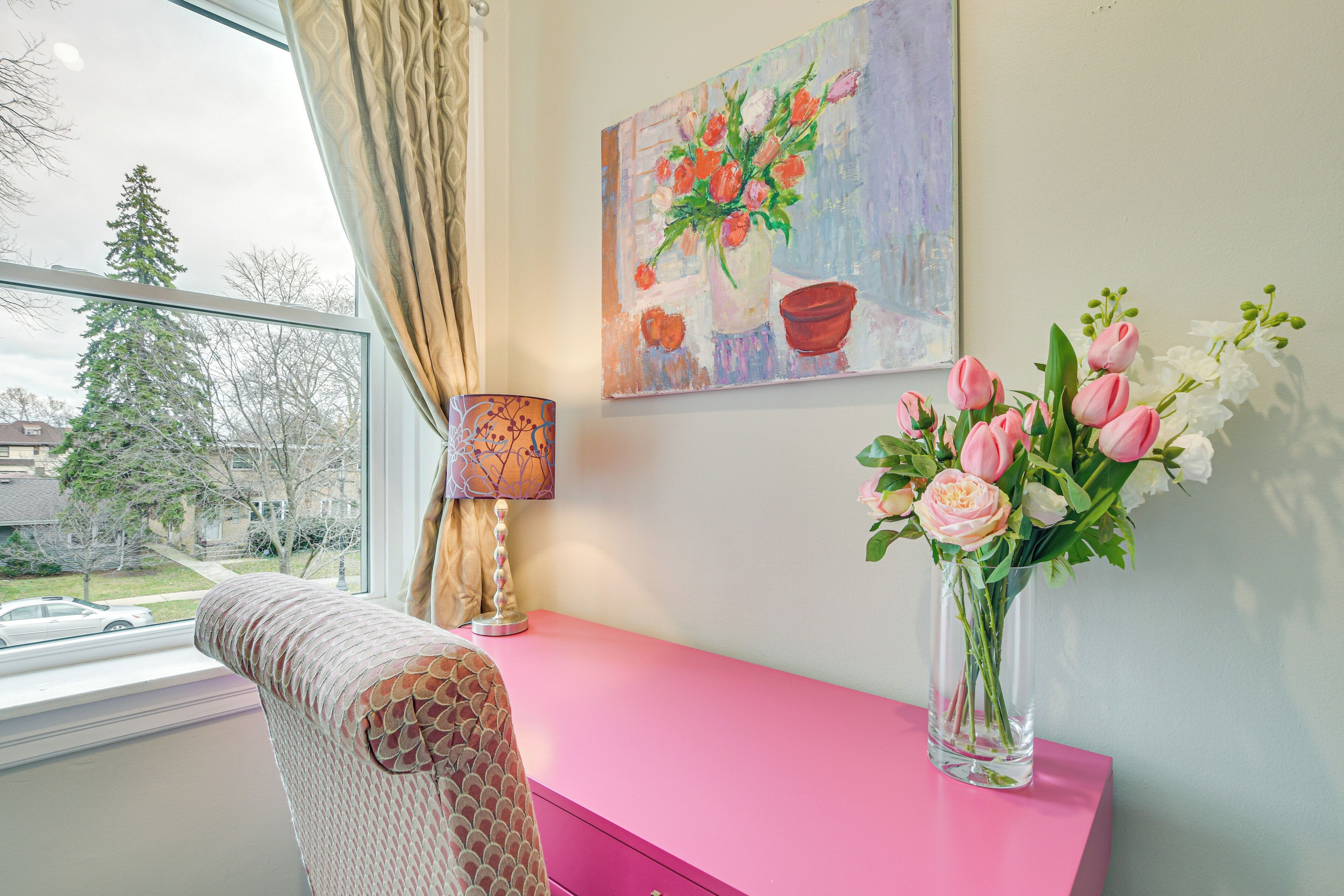 A room with a pink desk, a vase of flowers, a painting on the wall, and a window with a view of trees and a car.