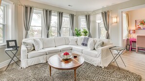 A living room with a white sectional sofa, a round wooden coffee table, and a large window with curtains. - Family Apartment w/ Deck + Yard Near Northwestern (Evanston)