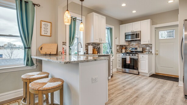 A modern kitchen with white cabinets, a central island with stools, stainless steel appliances, and a view of the outside. - Family Apartment w/ Deck + Yard Near Northwestern (Evanston)