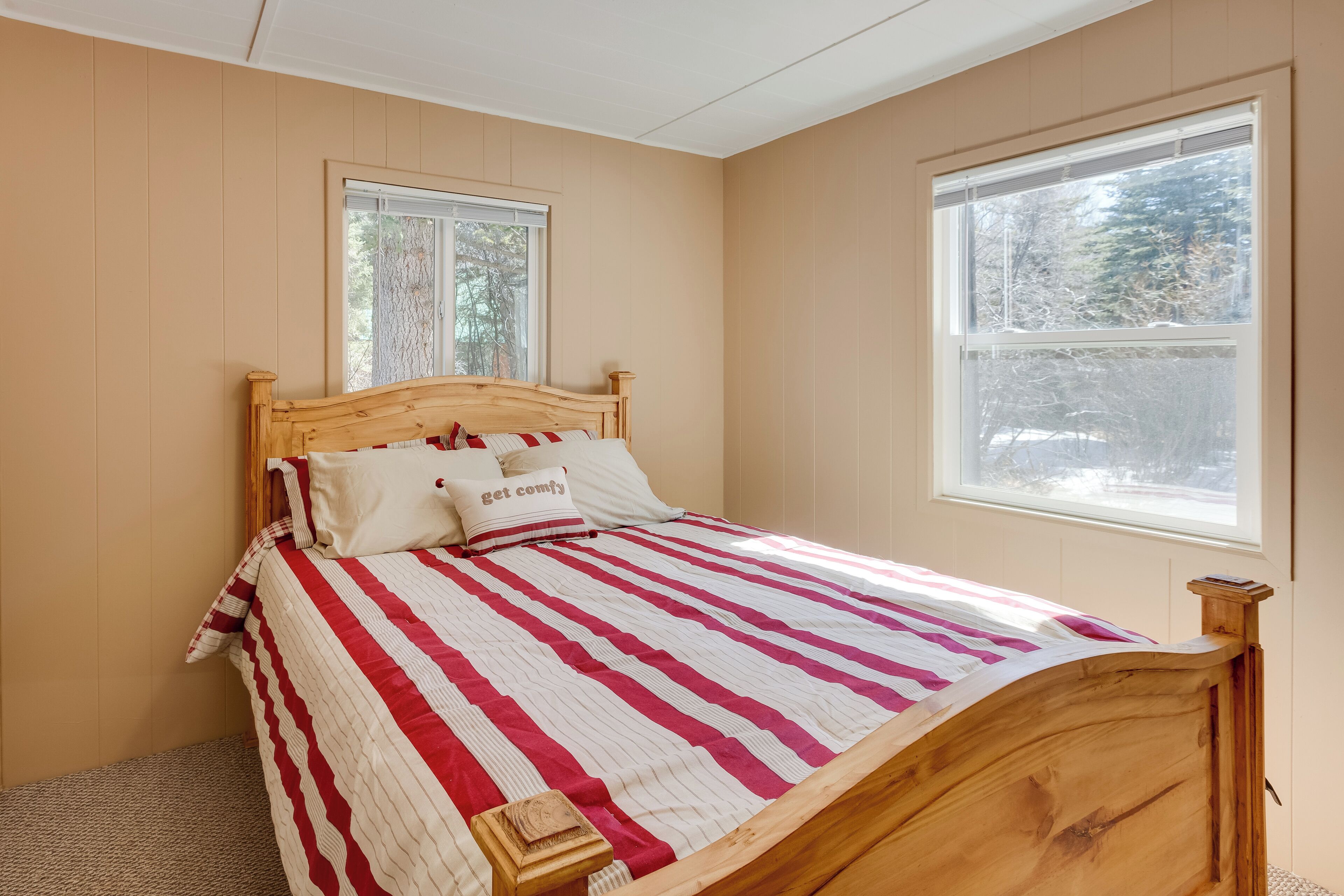 Ein Schlafzimmer mit einem hölzernen Bett, gestreifter Bettwäsche, zwei Fenstern und Blick auf Bäume.