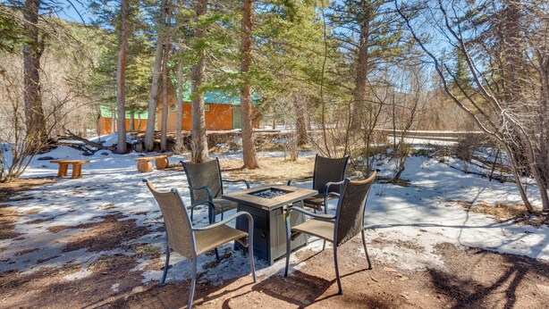 Une terrasse avec une table et des chaises, entourée d’arbres, et une cabane en arrière-plan.