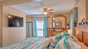 Une chambre à coucher comprenant un lit, un ventilateur de plafond, un téléviseur fixé au mur, une commode en bois, un miroir et une fenêtre avec des rideaux.