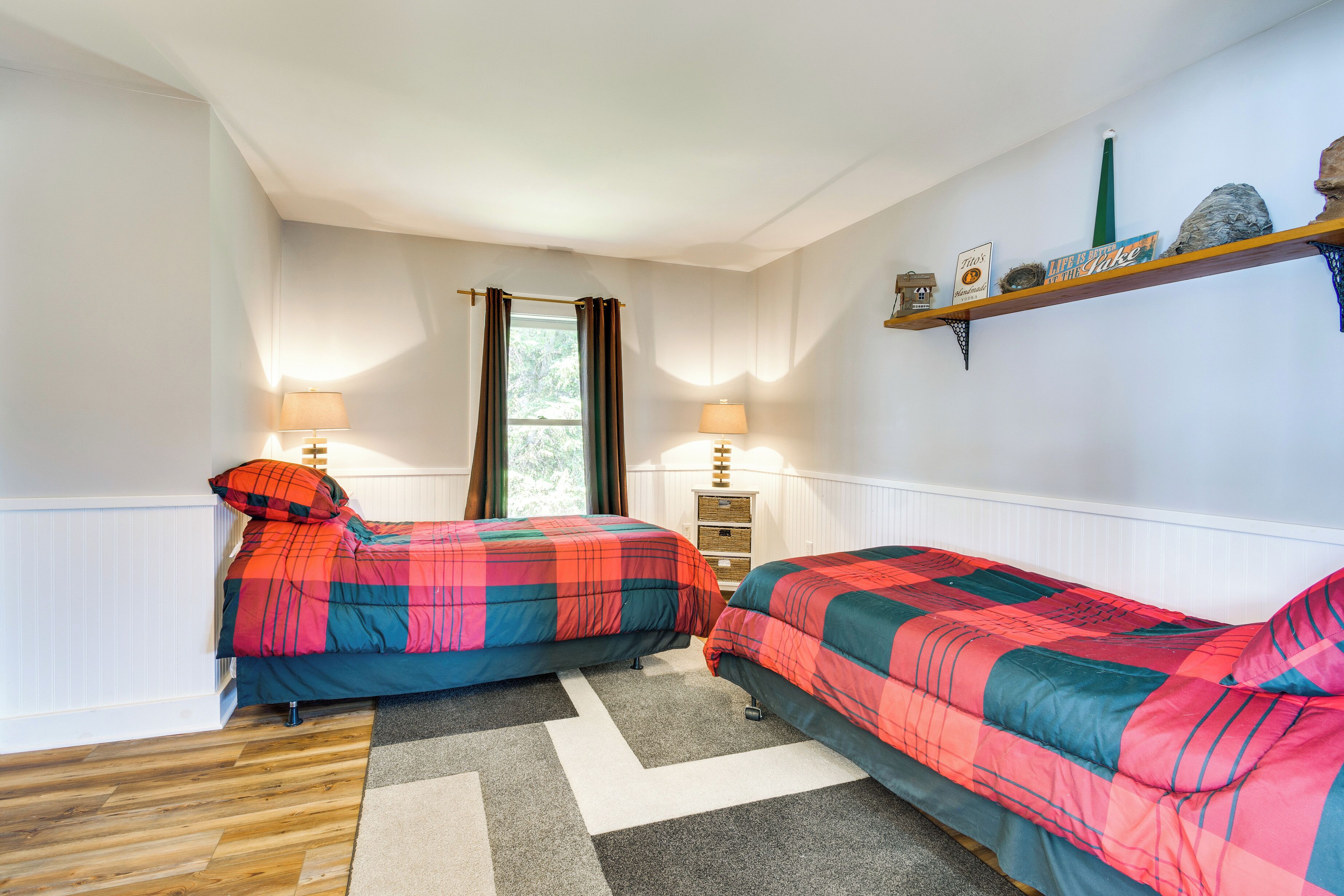 Un dormitorio con dos camas, una ventana con cortinas, una estantería con objetos decorativos y una lámpara en cada cama.