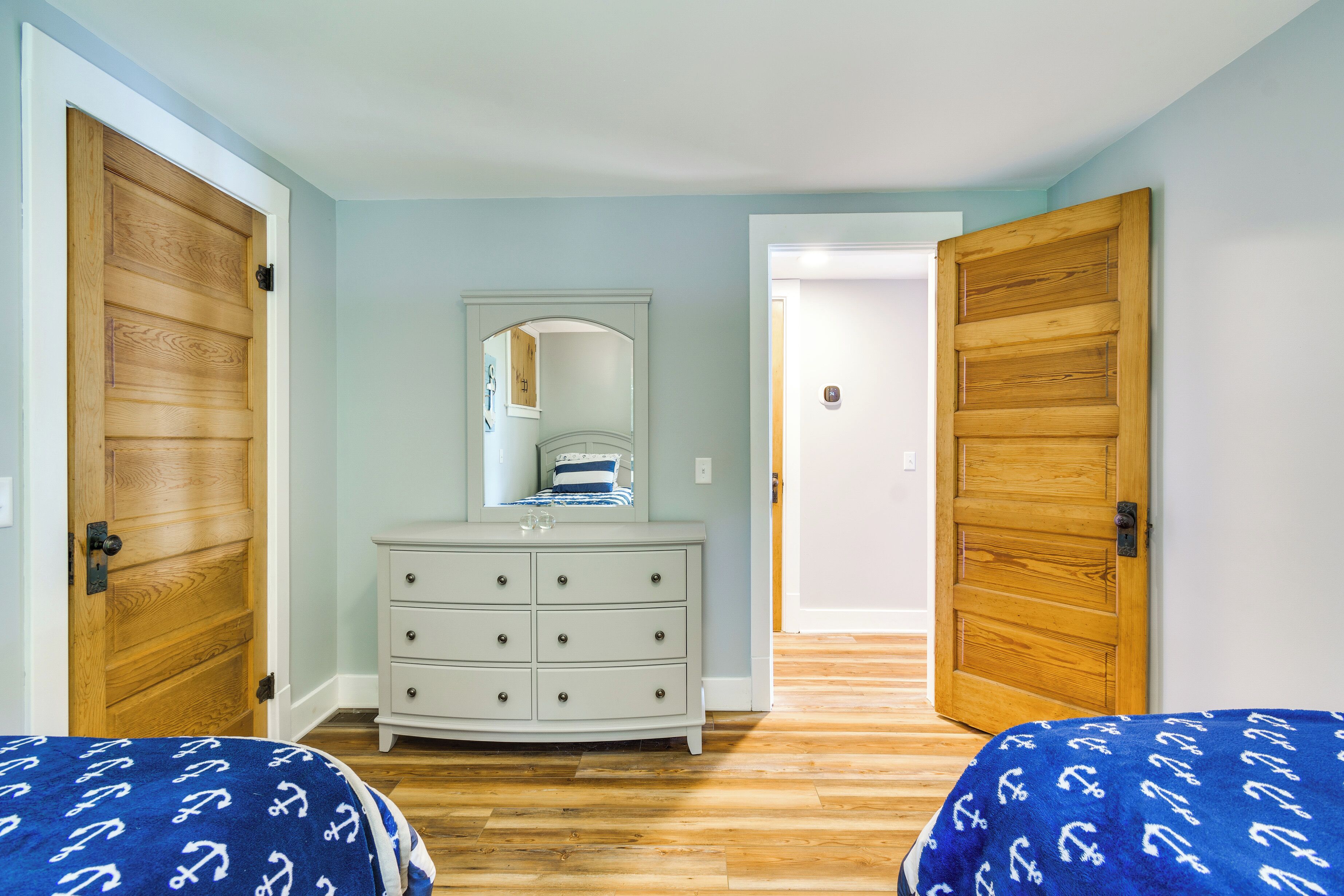 A bedroom with two wooden doors, a white dresser, a mirror, and two beds with blue bedding.