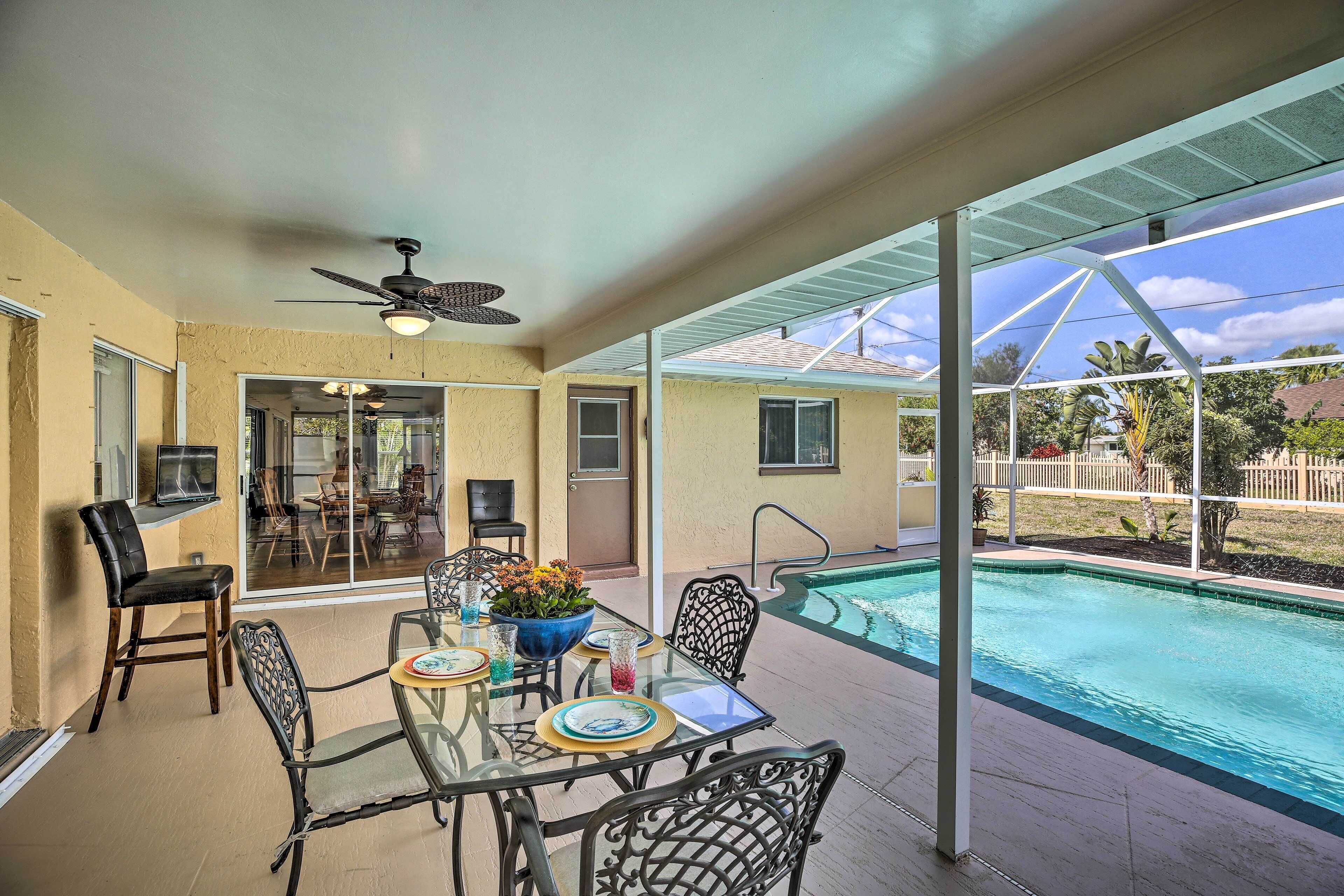 An outdoor patio with a glass table and chairs, a ceiling fan, and a view of a swimming pool.