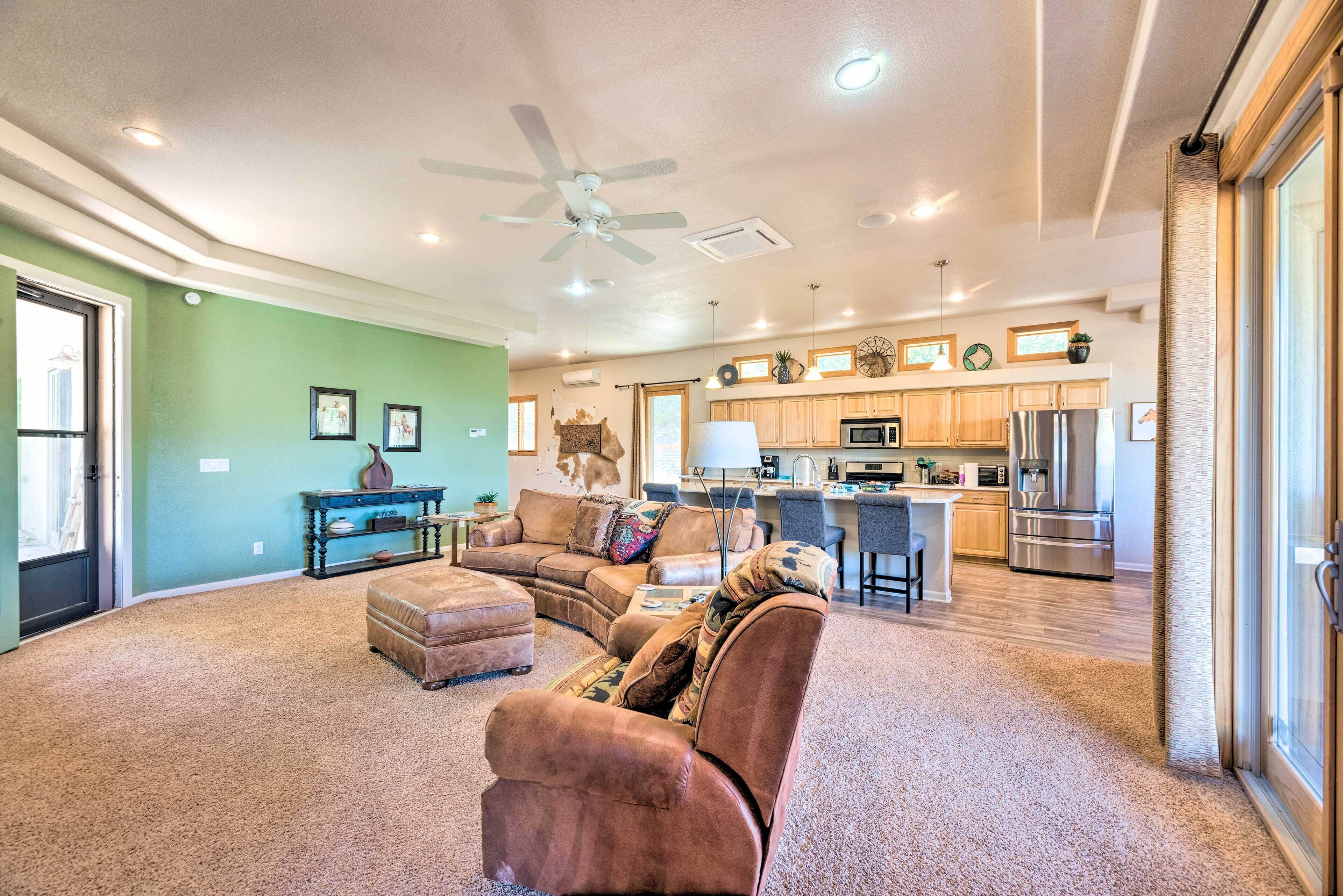 A spacious living room with a sectional sofa, ottoman, and a ceiling fan. A kitchen with modern appliances and a dining area is visible in the background.