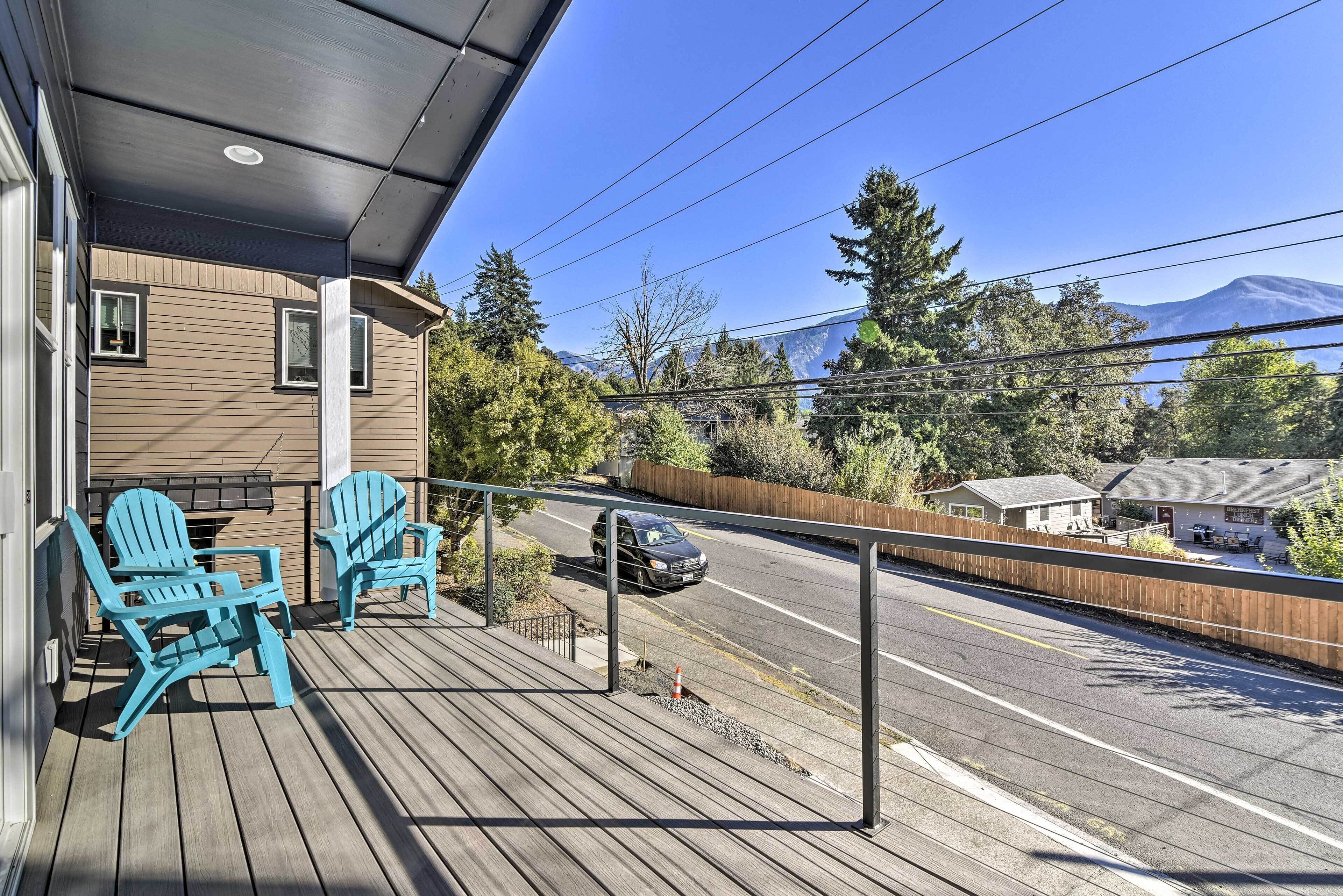 A wooden deck with two blue Adirondack chairs, a covered area, and a view of a road and mountains.