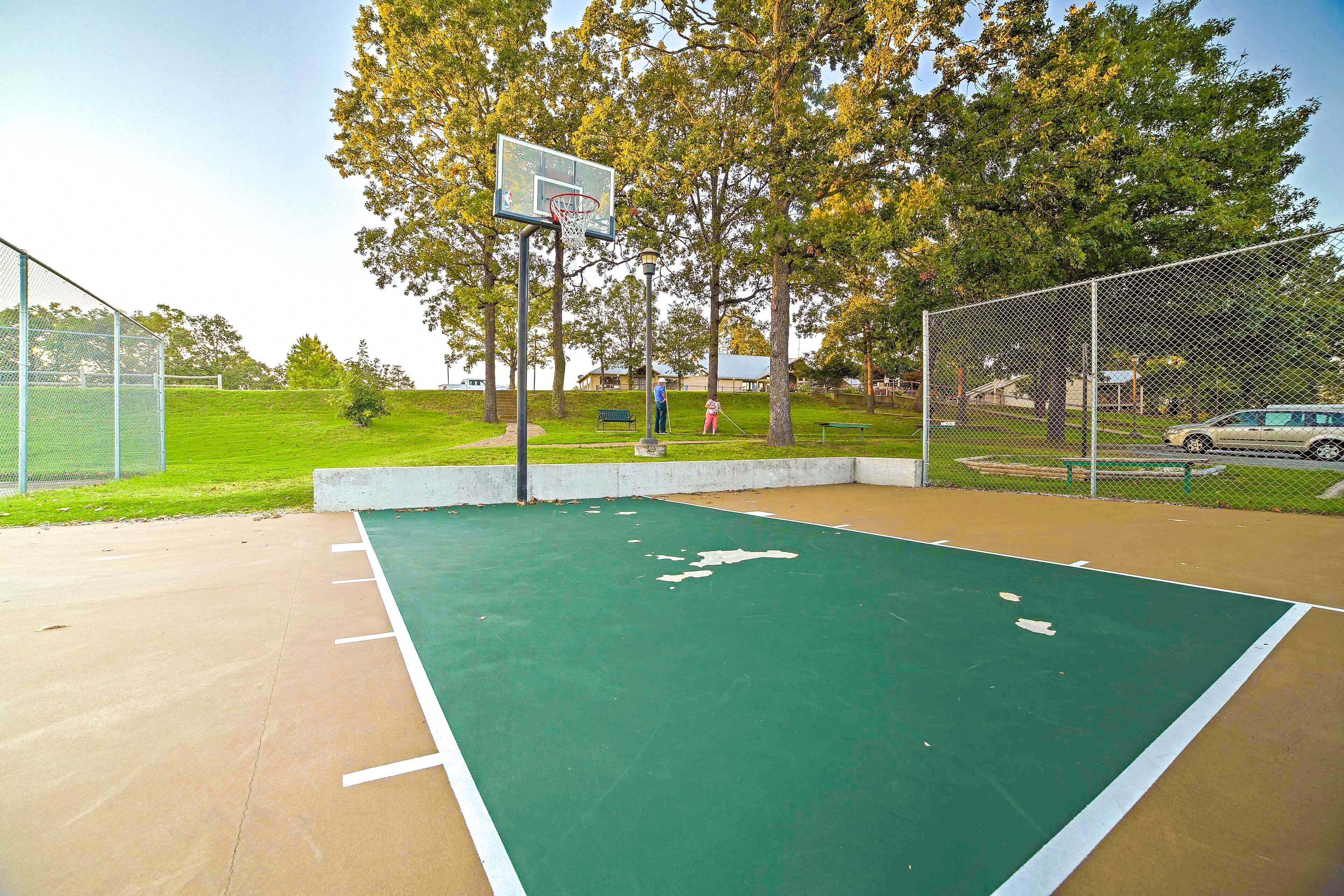 Un terrain de basket en plein air, doté d’une surface verte, entouré d’une clôture en treillis métallique, d’arbres et d’une voiture garée à proximité.