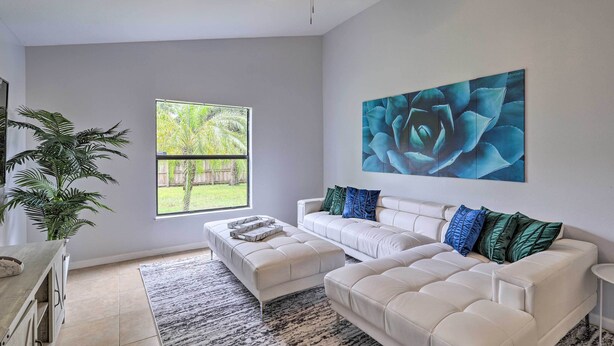 A spacious living room with a large window, a ceiling fan, a white sofa set, and a large abstract painting. - Charming Palm Bay Retreat w/ Spacious Yard! (Palm Bay)