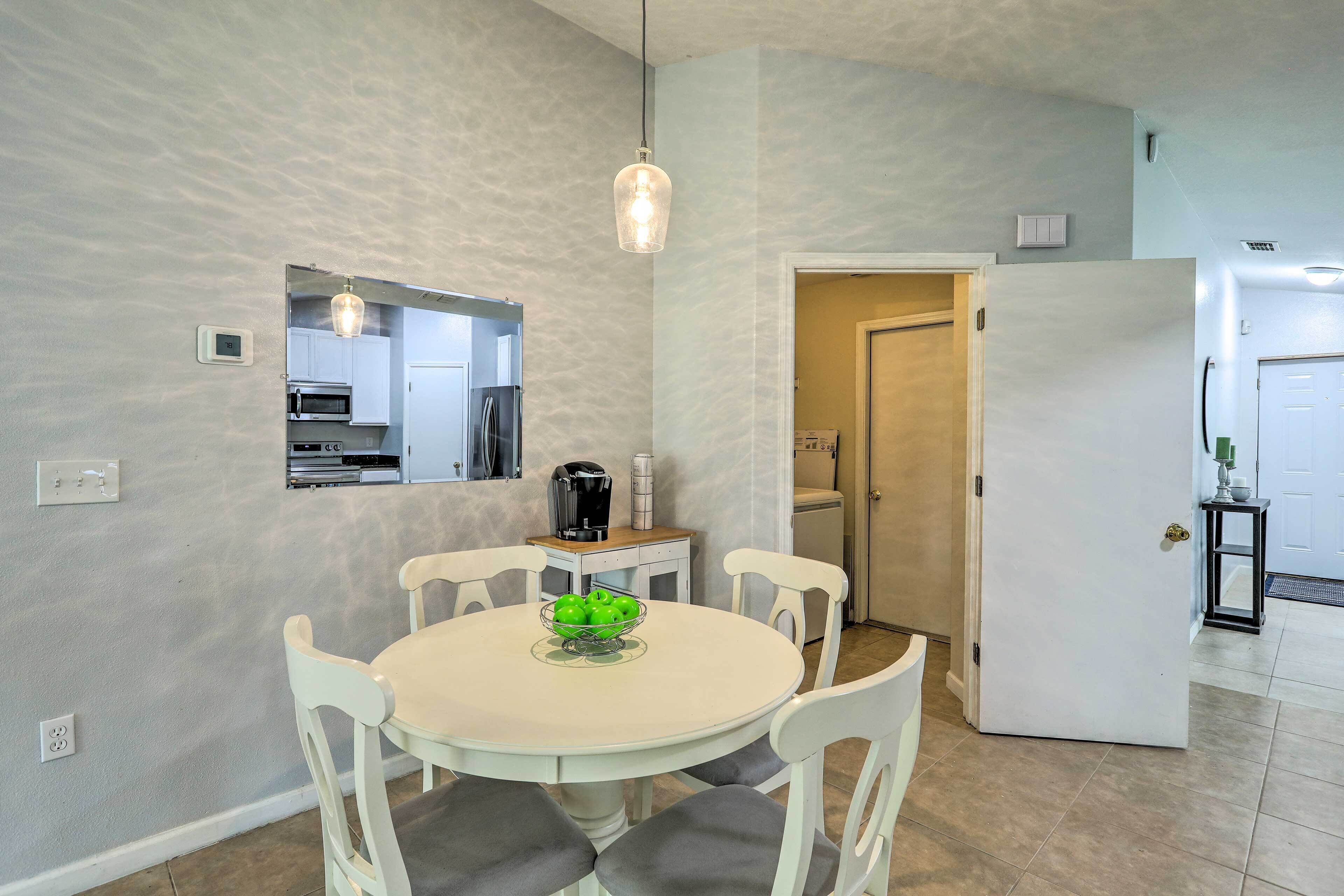 A dining area with a round table, white chairs, and a bowl of green apples. There is a kitchen in the background with a microwave and a coffee maker.