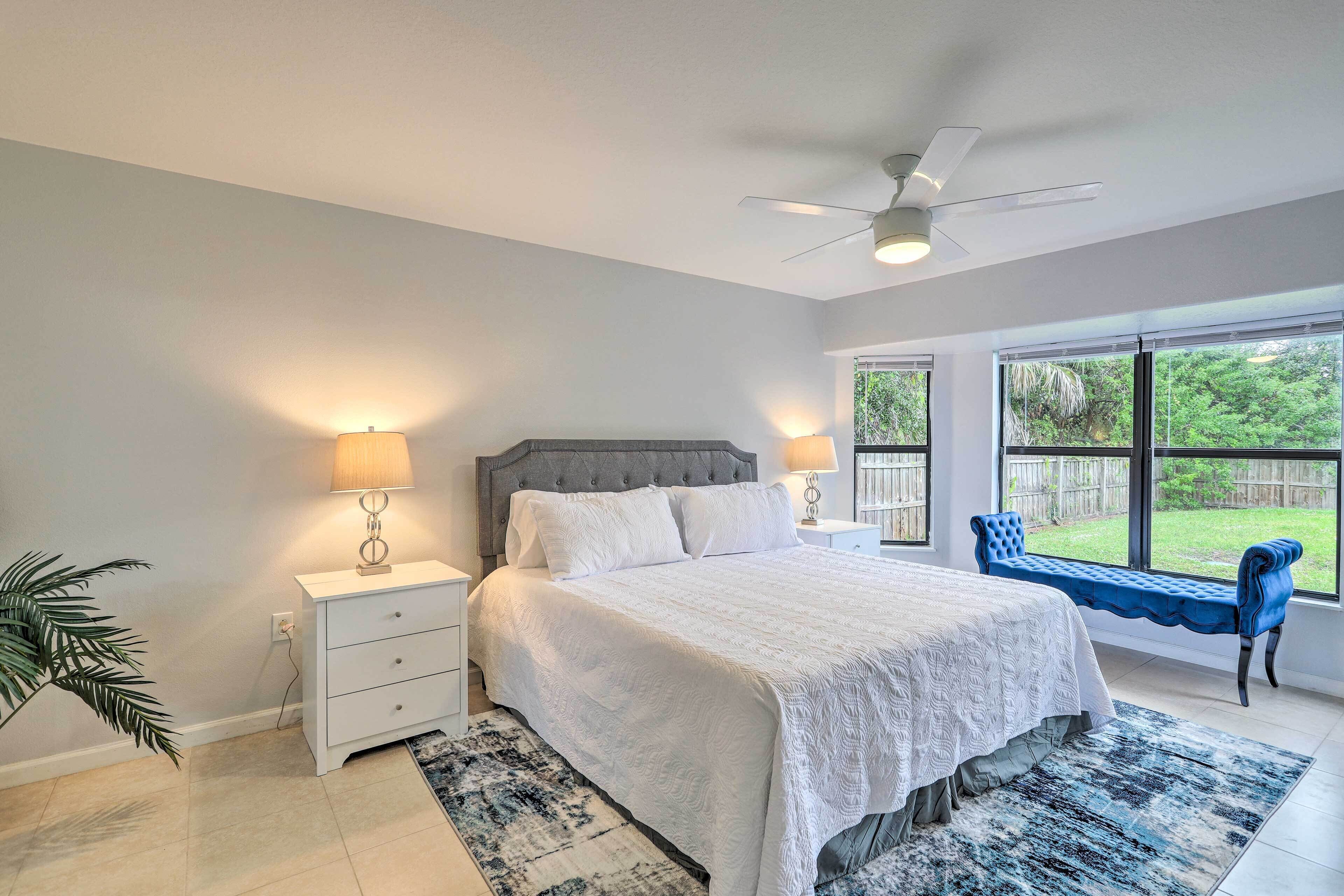A bedroom with a bed, bedside lamps, a ceiling fan, a window with a view of greenery, and a bench by the window.