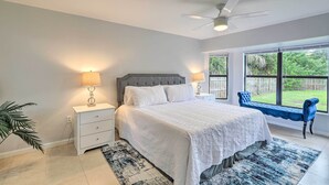 A bedroom with a bed, bedside lamps, a ceiling fan, a window with a view of greenery, and a bench by the window. - Charming Palm Bay Retreat w/ Spacious Yard! (Palm Bay)