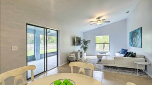 A spacious living room with a dining area, a ceiling fan, a large window, and a sliding glass door leading to a patio. - Charming Palm Bay Retreat w/ Spacious Yard! (Palm Bay)