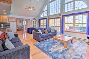 A spacious living room with a gray sofa, a wooden coffee table, and a blue rug. There are large windows with blue curtains, a ceiling fan, and a kitchen area in the background.