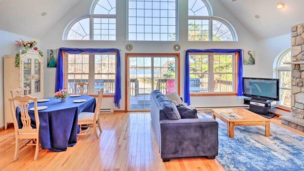 A spacious living room with a high ceiling, large windows, a fireplace, a television, and a dining area with a table and chairs. - Near Hiking: Lakefront Albrightsville Home w/ Deck (Albrightsville)