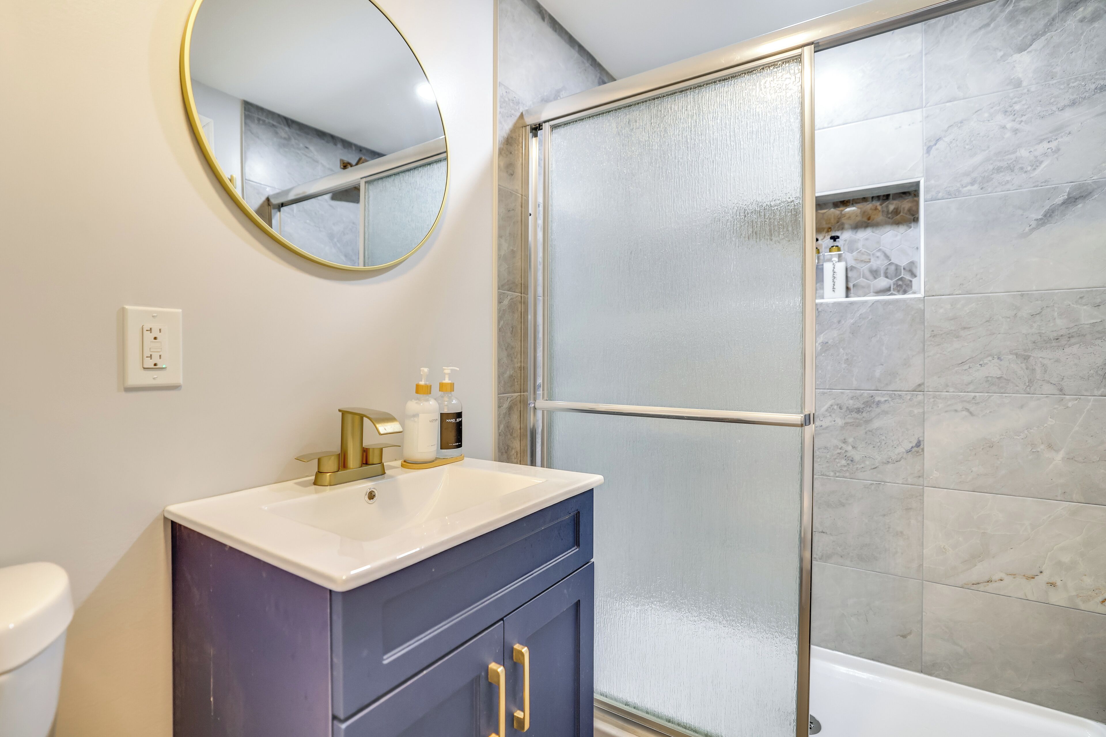 A bathroom with a white sink, dark blue vanity, gold handles, a round mirror, and a glass shower enclosure.