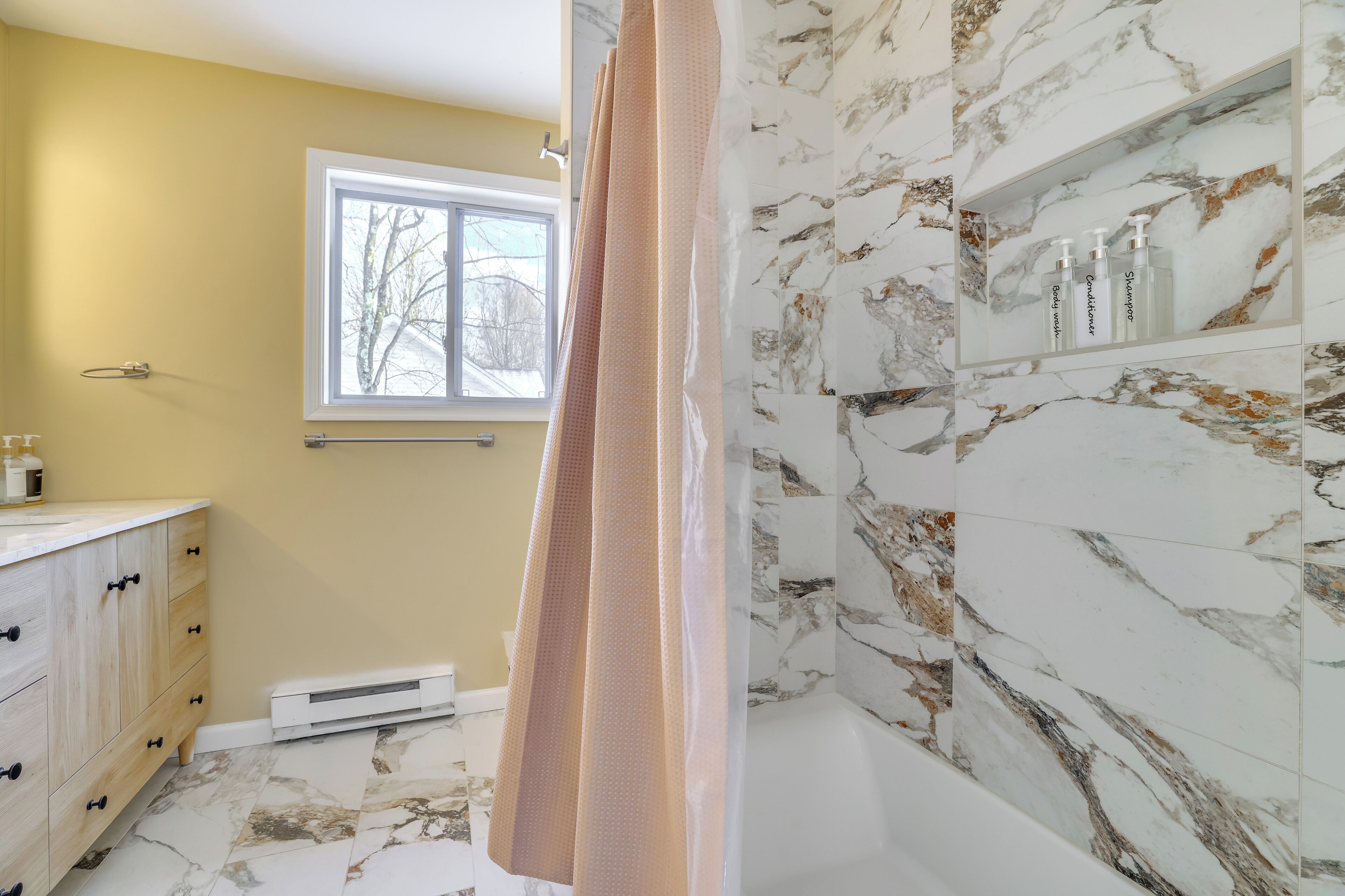 A bathroom with a marble wall, a window, a vanity with drawers, and a shower curtain.