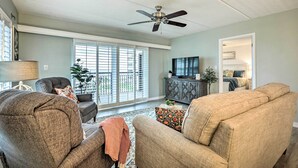 A living room with a ceiling fan, a large window with blinds, and a television on a cabinet. - Charming Ormond Beach Condo w/ Ocean Views! (Ormond Beach)