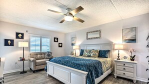 A bedroom with a bed, a nightstand, a chair, a ceiling fan, and a window with blinds. - Charming Ormond Beach Condo w/ Ocean Views! (Ormond Beach)