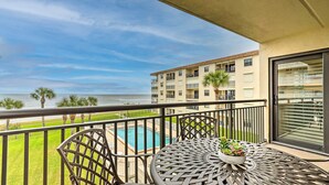 A balcony with a view of a pool, palm trees, and a building across the water. - Charming Ormond Beach Condo w/ Ocean Views! (Ormond Beach)