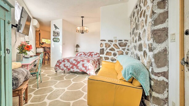 A cozy room with a bed, a yellow armchair, a desk with a computer, and a stone wall.