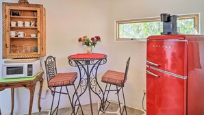 A compact kitchen with a red Galanz refrigerator, a wooden cabinet with dishes, a small table with chairs, and a window with a view of trees.