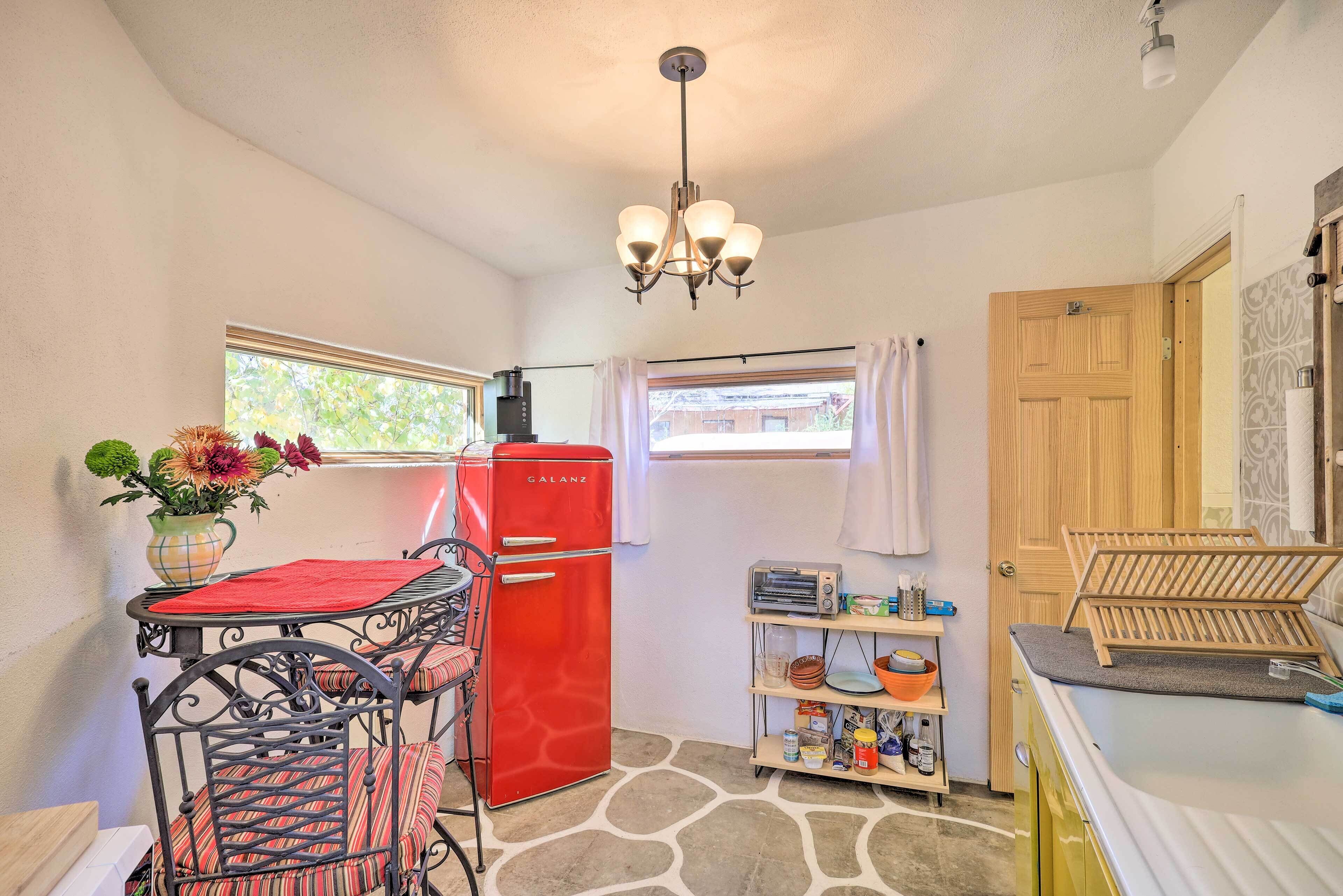 A compact kitchen with a red refrigerator, a dining table with chairs, a small shelf with kitchen items, and a window with curtains.