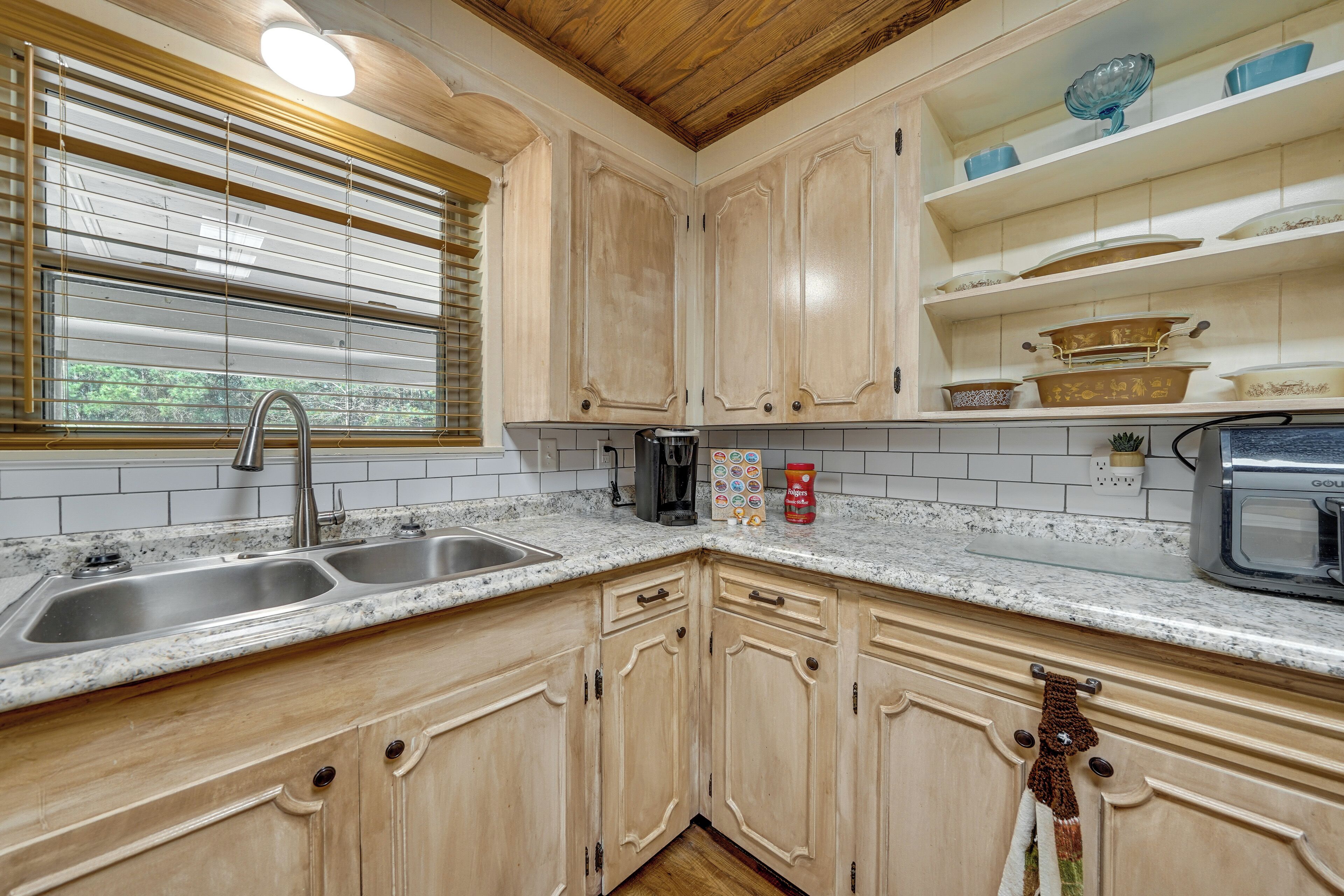 A kitchen with wooden cabinets, a double sink, a window with blinds, and a microwave.