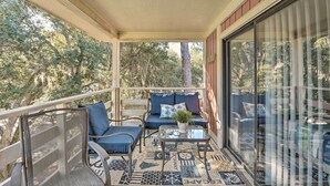 Un balcón techado con mobiliario exterior, una mesa de vidrio y vistas a árboles.
