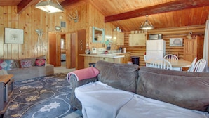Ein gemütliches Blockhaus-Interieur mit Wohnbereich, ausgestattet mit einer Couch, einem Couchtisch und einem Teppich. Eine Küchenzeile bietet einen Kühlschrank, eine Mikrowelle und einen Esstisch mit Stühlen.