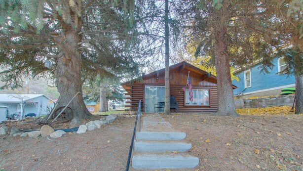Une cabane aux murs en bois, un escalier en béton menant à l’entrée, et une vue sur une maison bleue en arrière-plan.