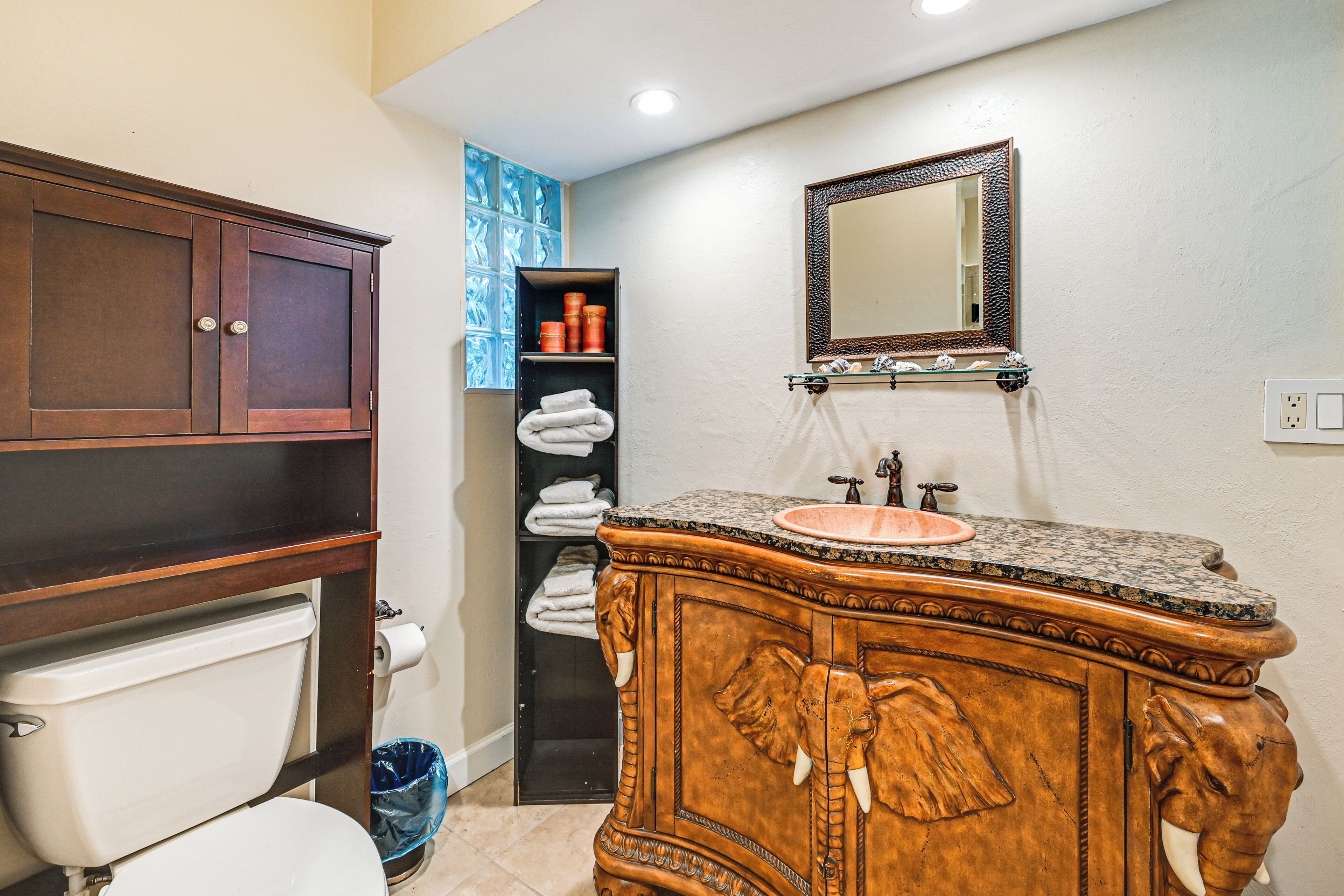 A bathroom with a wooden vanity, a sink with a marble countertop, a toilet, and a shelf with towels.