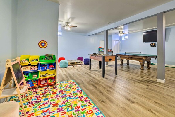 Une pièce avec un plancher en bois, un tapis coloré représentant l’alphabet, une table de baby-foot, une table de ping-pong et une bibliothèque remplie de jouets.