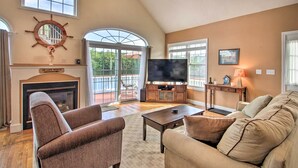 A living room with a fireplace, a television, and a large window overlooking a balcony. - Waterside Haven on Milton Pond w/ Deck & Boat Dock (Milton)