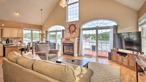 A spacious living room with a fireplace, a television, and a large window overlooking a deck. - Waterside Haven on Milton Pond w/ Deck & Boat Dock (Milton)