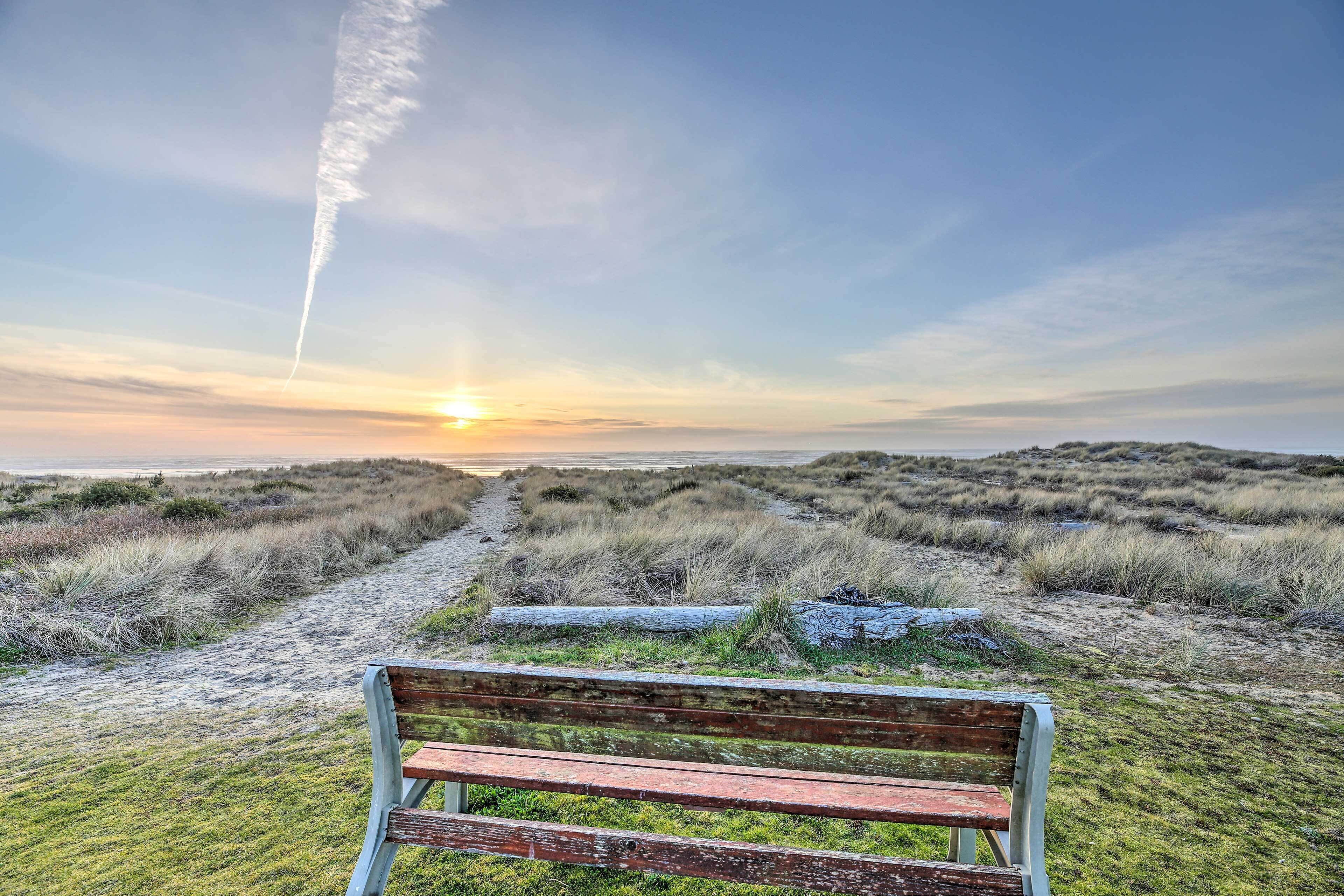 A bench in a natural setting with a path leading to the ocean and a sunset in the background.