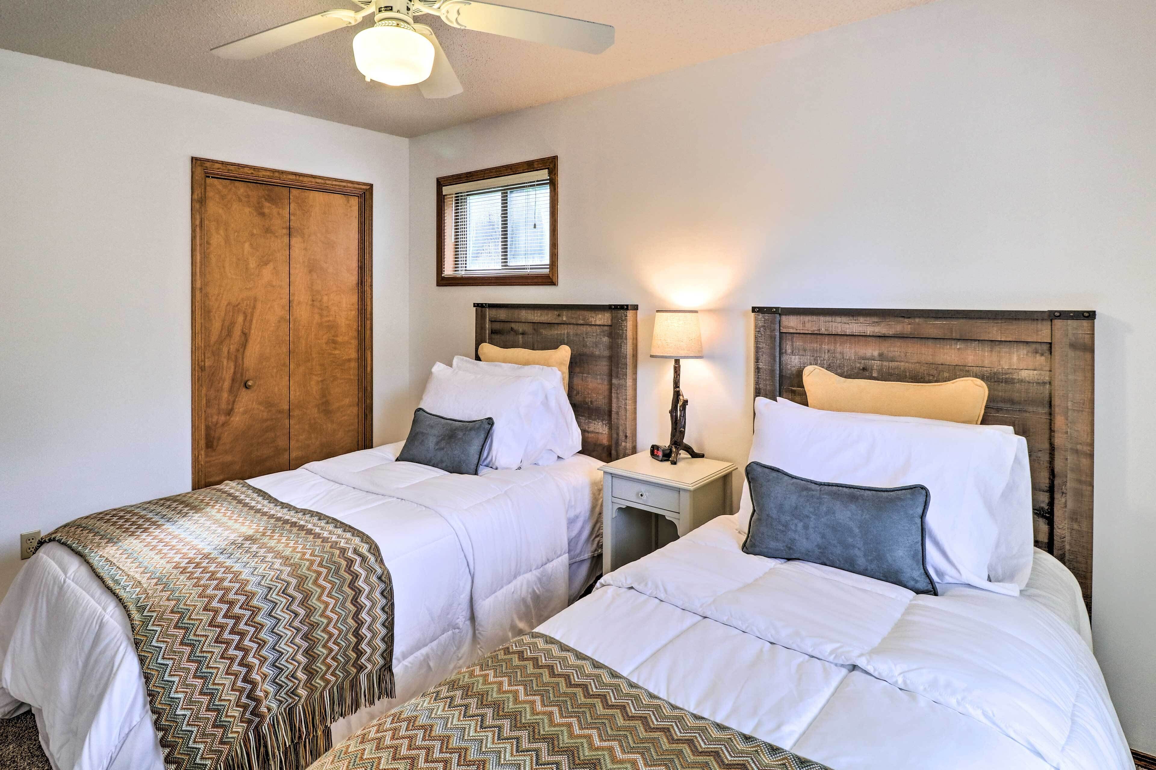 Une chambre d’hôtel avec deux lits, des têtes de lit en bois, une table de chevet avec une lampe, un ventilateur de plafond et une fenêtre avec des stores.