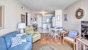 A living room with a sofa, armchair, and dining table. There is a kitchen in the background with cabinets and a refrigerator. - North Myrtle Beach Condo w/ Stunning Ocean Views! (North Myrtle Beach)